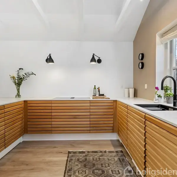 Køkken med træbeklædte skabe, sort armatur og to vægmonterede lamper. En blomstervase står på bordet, og et vindue lader naturligt lys ind i rummet. Tæppe på gulvet.