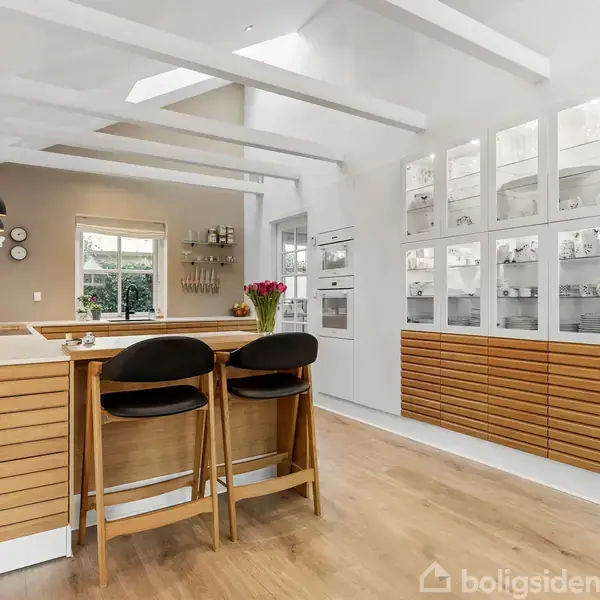 To høje barstole står ved køkkenbordet i et lyst, moderne køkken med træelementer. Vaser med blomster pynter bordet. Et vindue og væghylder ses i baggrunden.