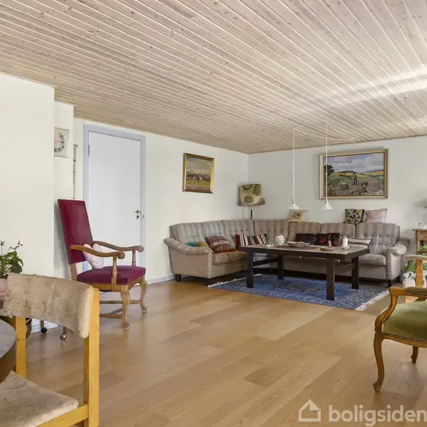 Stue med møbler, hvor en grå hjørnesofa står ved siden af et brunt sofabord på et blåt tæppe. Trægulv og træbeklædt loft skaber en varm atmosfære.