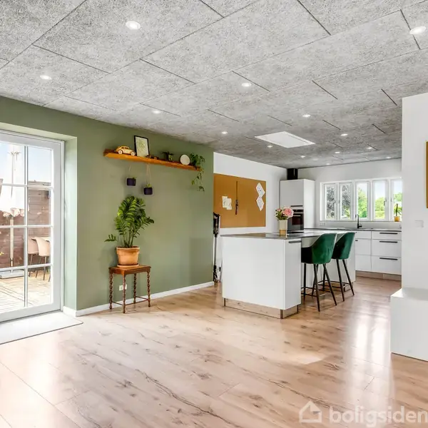 Lyst køkkenområde med hvidt interiør og køkkenø med grønne barstole. Store vinduer åbner ud mod terrasse, grøn plante og hylde på malet væg. Træfarvet gulv.