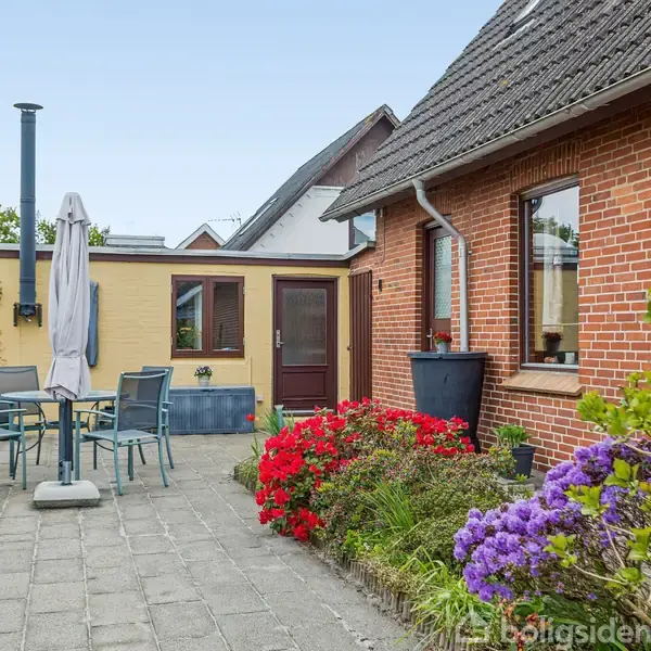 Rundt havebord med stole og en parasol står på en flisebelagt terrasse ved siden af et rødt murstenshus, omgivet af blomsterbede med farverige blomster og grønne planter.