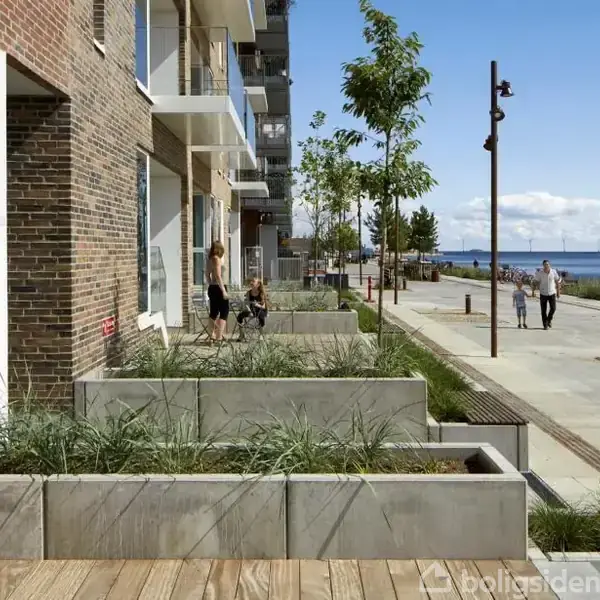 Bygning med røde mursten har store plantekasser ude foran. Flere mennesker går langs en havnepromenade med udsigt til vandet, og nummer 9 er synligt på døren.