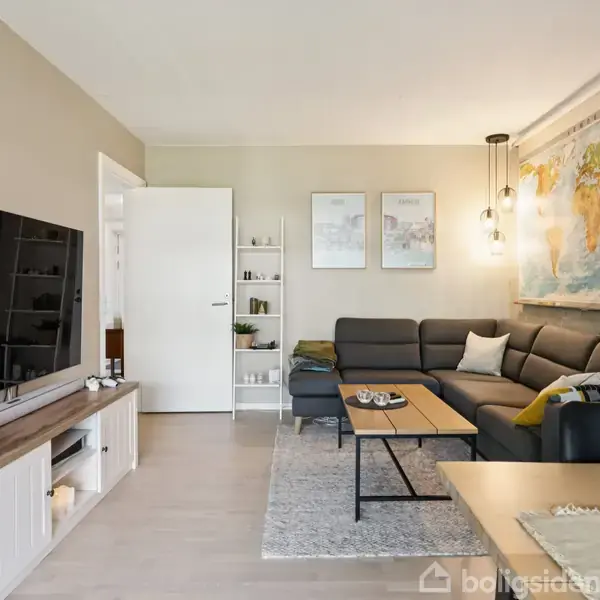 En stue med en stor grå hjørnesofa ved en væg med et verdenskort. Et træbord foran sofaen. Fladskærms-TV og reol til venstre. Bucharer og tre billeder på væggen.