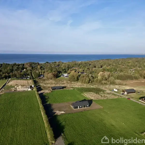 Et hus står på en grøn mark, omgivet af marker og træer. I baggrunden ses en bred havudsigt under en blå himmel.