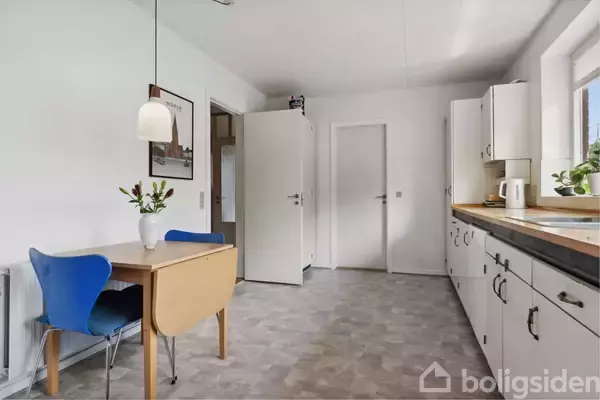 Et køkken med et spisebord og en blå stol står ved en væg. Bordet har en vase med blomster. Hvide skabe og apparater fylder rummet, der er klart oplyst af dagslys.