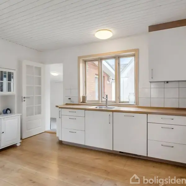 Køkken med lyse træbordplader og hvide skabe; lys strømmer gennem et stort vindue over vasken; trægulv og hvidt loft; dør fører til en anden del af huset.