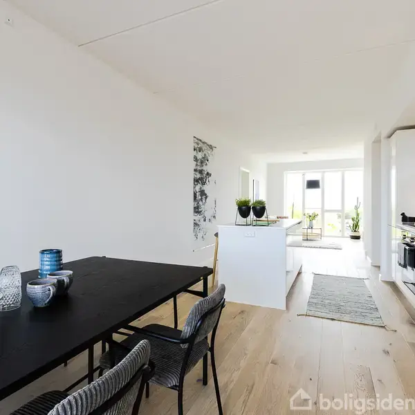 Et sort spisebord er dækket med service, omgivet af stole, i et moderne køkken-alrum med hvide skabe og trægulv. Store vinduer bagest lader lys strømme ind.