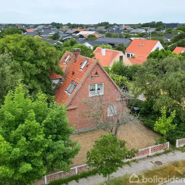 En rød murstensvilla med skråt tag står omgivet af træer i et parcelhuskvarter. Omkringliggende haver og huse med orangerøde tage ses i det grønne landskab.