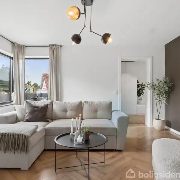 Sofa med puder står i stue, omkranset af store vinduer med gardiner. Et rundt sofabord med lys og pynt. Trægulv, kunst på væggen og planter som dekoration.