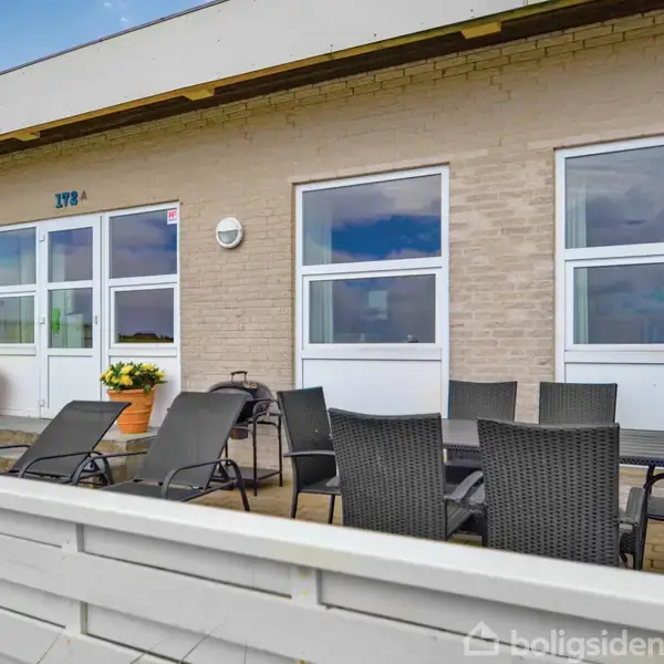 Hus med lys murstensfacade har en terrasse med sorte havemøbler og liggestole. Der er to potteplanter ved indgangen, markeret med nummeret "192 A".