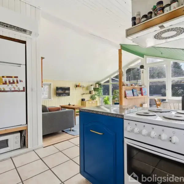 En åben køkkenløsning med en komfur ø med blå skabe, omgivet af en stue med store vinduer og en sofa i et lyst rum. Der er planter og hylder med krydderier.