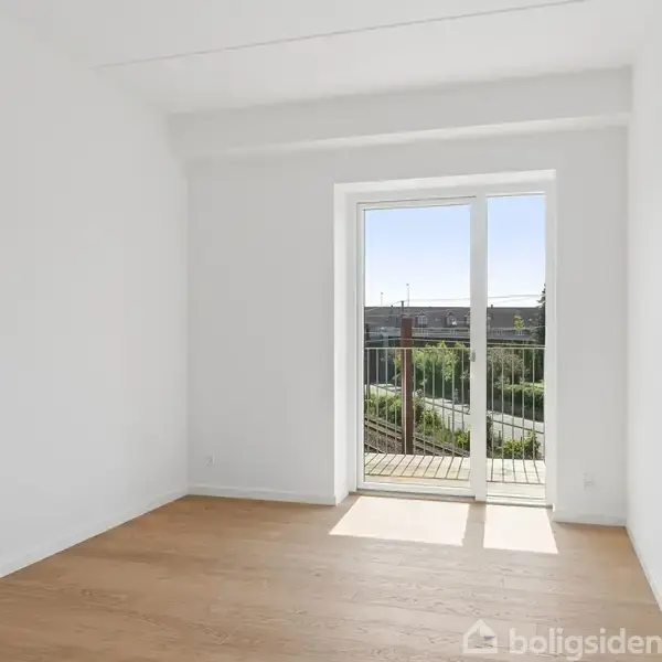 Et lyst, tomt rum med trægulv og en stor glasdør åbner ud til en altan. Sollys strømmer ind og oplyser rummet, der er hvidmalet med en klar himmel udenfor.