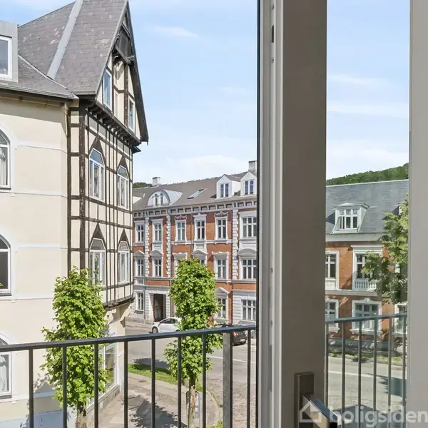 En udsigt fra en balkon viser en gade med klassiske bygninger i flere etager og træer i vejkanten, i en stille bydel med blå himmel.