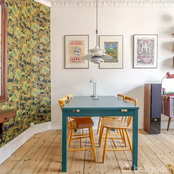 En grøn spisebord står i et farverigt rum med tapet, omgivet af fire træstole. Væggene har kunstplakater og en bogreol. Trægulvet skaber en varm atmosfære.