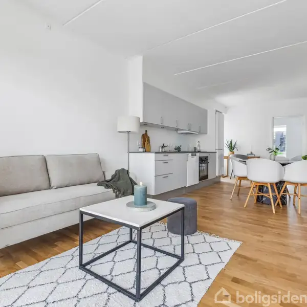 Grå sofa står ved siden af et sofabord med lys på en hvidt tæppe, i en moderne stue-køkken kombination med træparketgulve og spisebord i baggrunden.