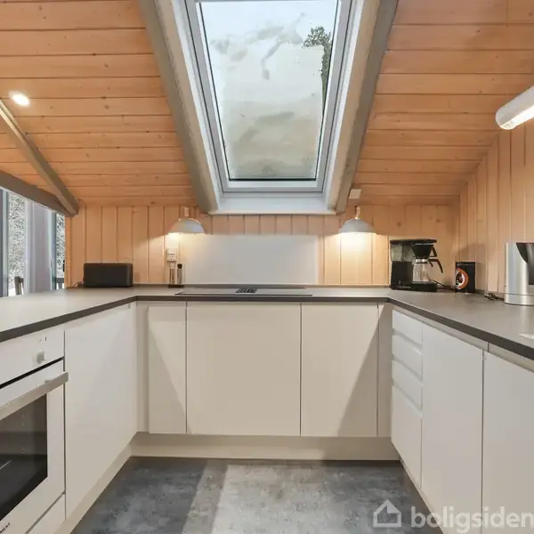 Et moderne køkken med lyse skabe og indbygget ovn. Ovenlys vindue og træloft skaber lys atmosfære. Kaffemaskine og elkedel står ved siden af vasken.