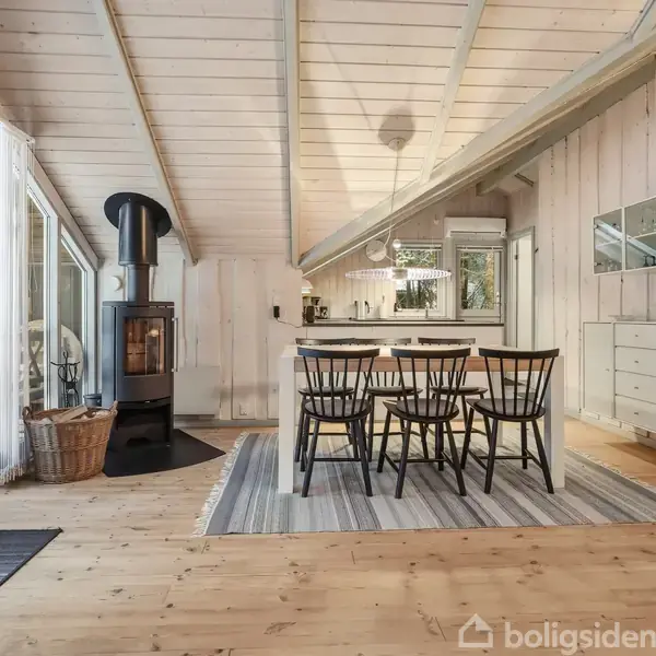 Brændeovn står tændt i en lys stue. Et spisebord med seks stole er placeret på et tæppe ved siden af store vinduer, der giver udsigt til udendørs omgivelser.