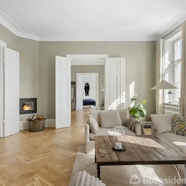 En stue med en beige sofa står ved et træbord. Sollys strømmer ind gennem store vinduer. I det tilstødende rum ses en seng. En plante og pejs fuldender scenen.
