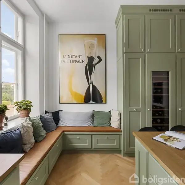 Bænk med grønne skabe og puder står ved et stort vindue. Ovenfor hænger en plakat med teksten "L'Instant Taittinger". Rummet har lyst trægulv og murstensvægge.