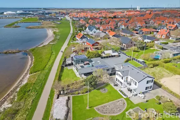 Huse langs en kyst med velplejede haver ligger ved siden af en sti. Byens røde tage strækker sig ud i baggrunden, mens en bugt og grønne områder omkranser scenen.
