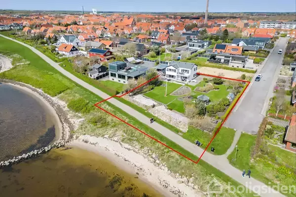 Hus set fra oven med stor have, ligger i et beboelsesområde nær kysten. Rød linje markerer grunden. Omkringliggende huse og veje danner et tæt fællesskab i det grønne miljø.