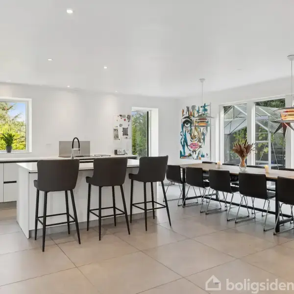 Køkken med hvidt interiør og stor ø formet ø med fire høje stole. Lang spisebord med otte stole ved siden af store vinduer, der viser en have. Indendørs planter og kunst på væggen.