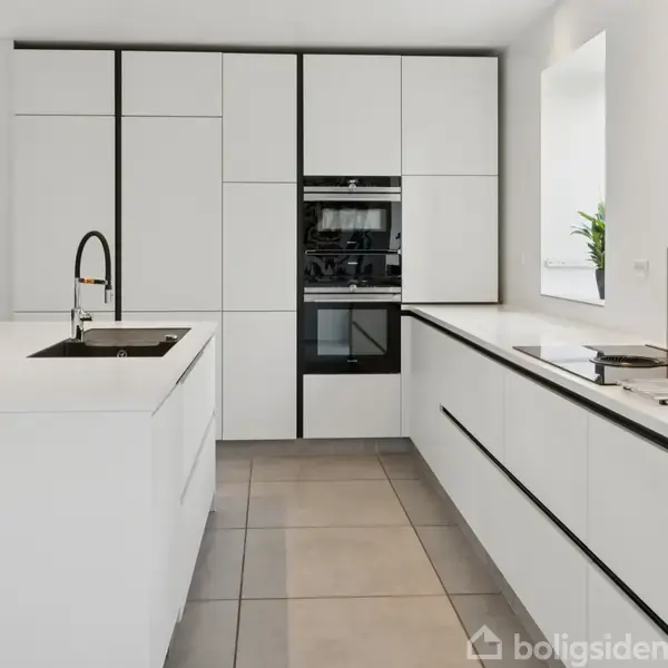 Hvidt køkken med indbygget ovn og skabsvæg; sort vandhane på køkkenø; store fliser på gulvet; vindue til højre med plante og masser af naturligt lys.