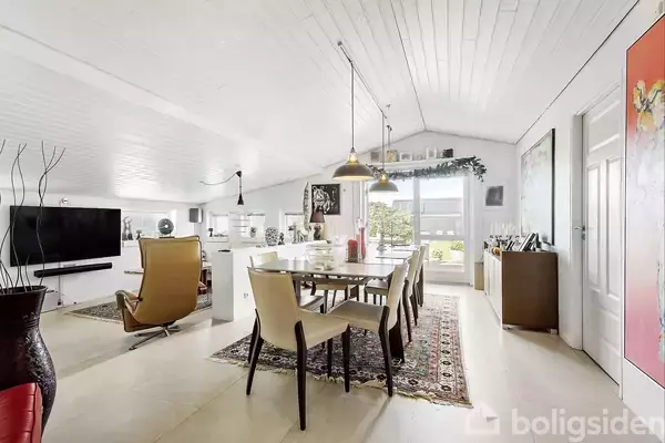 Et spisebord med stole står i et lyst stue-alrum. Baggrunden har store vinduer, der lukker naturligt lys ind. Rummet er indrettet med tæpper, planter og kunst på væggene.