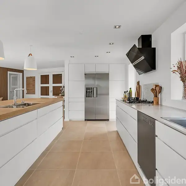 Et moderne køkken med hvide skabe og køkkenø. En stålvask og opvaskemaskine ses til højre. Store vinduer giver udsigt til en grøn have.