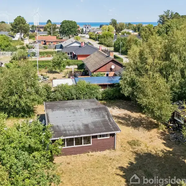 Et lille træhus står stille midt i en frodig have i et boligområde med udsigt over en fjern kyst. Græsplænen er tør, omgivet af forskellige træer og buske.