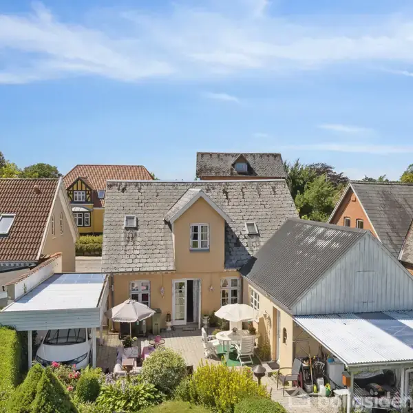 Et rækkehus med en gul facade har en terrasse med borde og parasoller. Huset er omgivet af naboer og velplejede haver under en blå himmel.