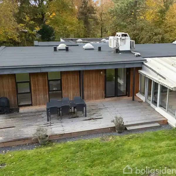 En moderne enfamiliehus med fladt tag står i en grøn have. En træterrasse med spisebord og stole er ved siden af en udestue. Omkring er der træer og græsplæne.