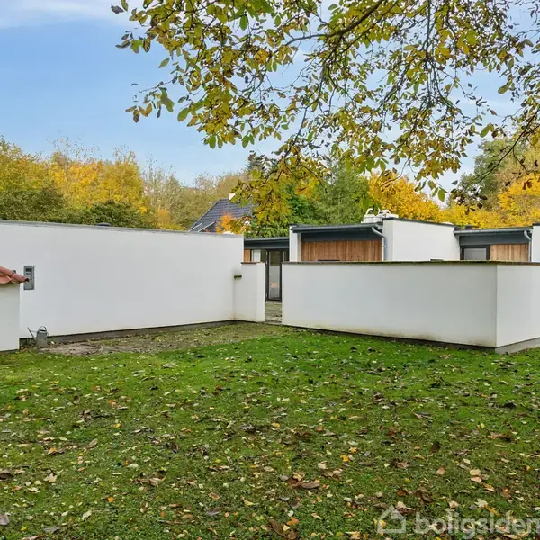 Moderne, lav, hvid bygning omgivet af grøn plæne og træer med efterårsblade. Bygningen har fladt tag og store vinduer, mens omgivelserne består af tæt løv og åbent græsareal.