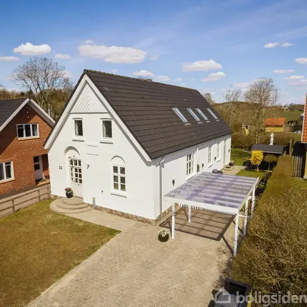Hvidt hus med sort tag står stille ved siden af to andre huse i en landsby. En carport er ved siden af huset, omgivet af hække og haver.