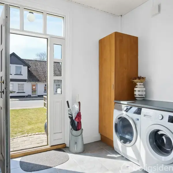 Vaskemaskine og tørretumbler står side om side i en lys entré med åbnet dør mod en forhave og udsigt til huse over vejen. En skabsenhed og paraplyholder ses ved siden.
