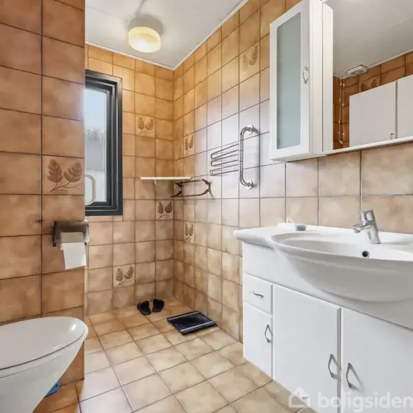 Toilet står ved siden af vasken i et badeværelse med brunlige fliser. Der er et vindue til venstre og skabe over vasken. Bruseområdet er i baggrunden.