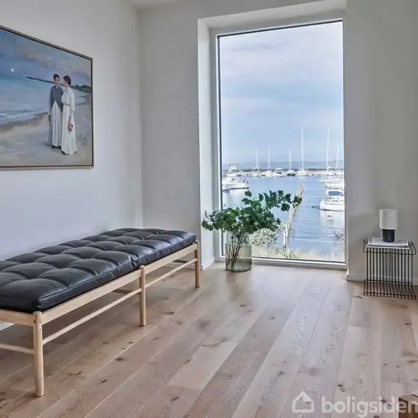 Bænk med sort hynde står ved vindue med havudsigt. Plakat af par på strand hænger på væg. Plante og lille bord med lysestage nær vinduet.