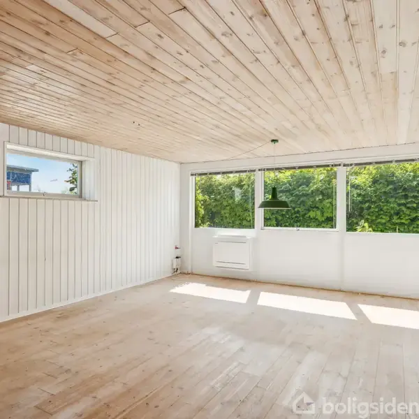 En lys og tom stue med trægulv og træloft. Sollys strømmer ind gennem store vinduer, der vender ud mod en grøn have. En enkelt lampe hænger fra loftet.