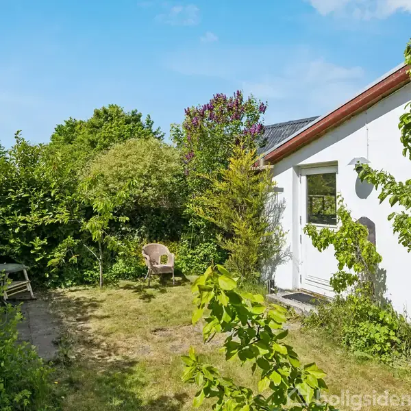 Hus omkranset af grøn have med en træbænk, planter og en sti. Hvid ydervæg og tag med ovenlys. Klar blå himmel i baggrunden.