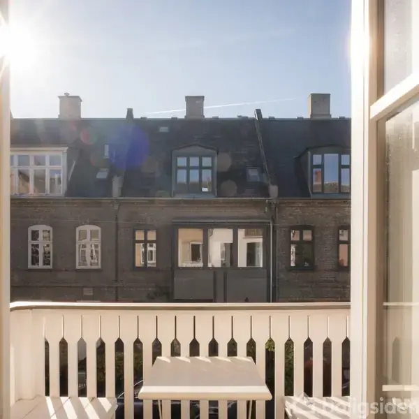 Balkon med hvidt rækværk, hvor et lille bord er placeret. Sollys skinner ind, og i baggrunden ses en række gamle bygninger under en klar blå himmel.