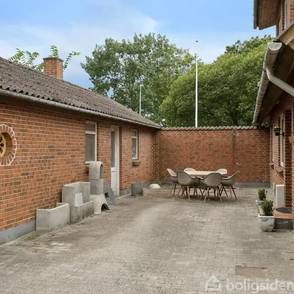 Et sæt bord og stole står stille i en brostensbelagt gårdhave, omgivet af røde murstensbygninger. Grønne træer er synlige i baggrunden.