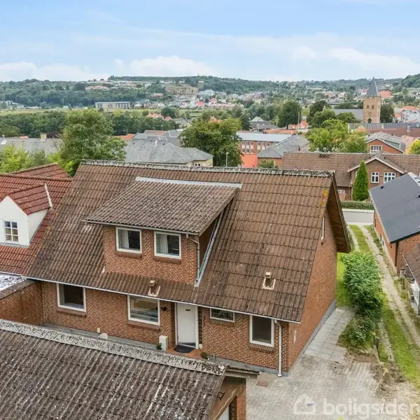 En rød murstensbygning med brunt tegltag står i en bymæssig setting med flere huse omkring og en kirke i baggrunden. Grønt landskab og træer ses i horisonten.