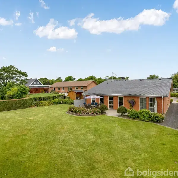 Rødt murstenshus står stille i en velholdt have med græsplæne og hække, omgivet af andre lignende huse under en klar blå himmel.