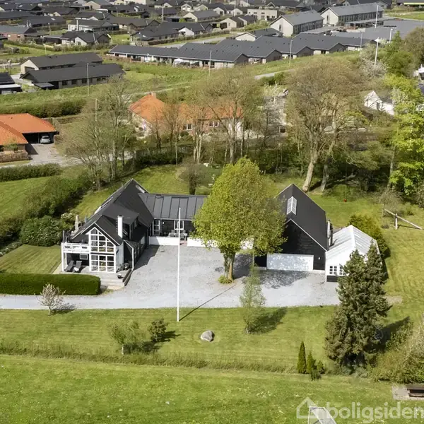 Hus placerer sig centralt på en stor grund med græs og træer, omgivet af villakvarter med flere huse i baggrunden og let sneklædte bjergtoppe.