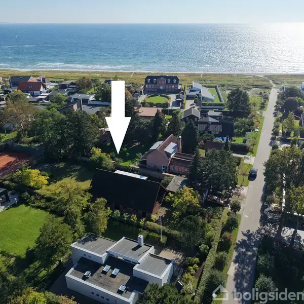 En pil peger på et hus i et landområde ved kysten. Huset er omringet af træer og haver, med en vej til højre og havet i baggrunden.