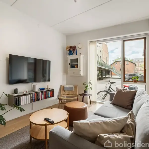 Sofa med puder står i en stue ved siden af et lille bord. Der hænger et fladskærms-tv på væggen over en hylde med bøger, en cykel står udenfor vinduet.