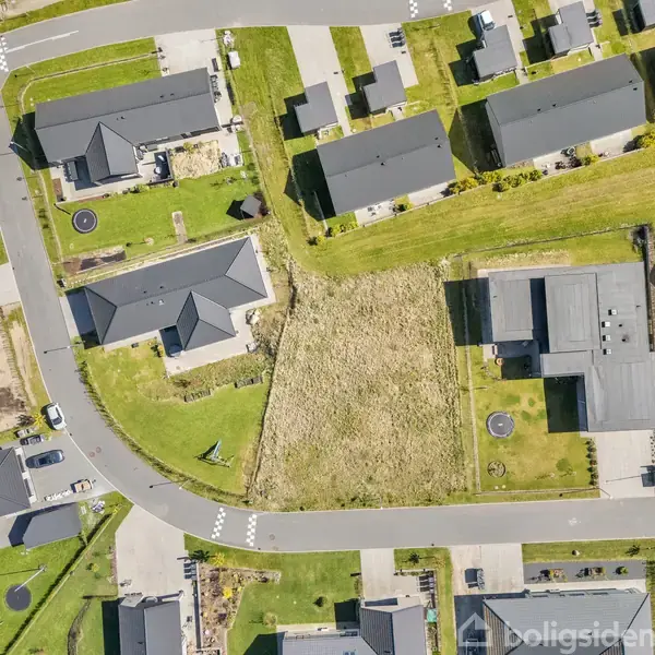 Luftfoto af boligområde med huse placeret omkring et trekantet, græsbevokset areal. Vejnettet snor sig rundt om området, og biler er parkeret langs vejene.