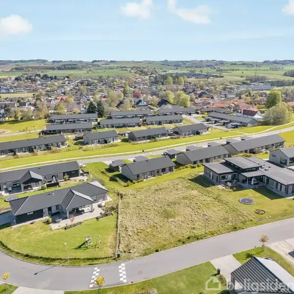 Boligområde med moderne huse placeret langs buede veje, omringet af grønne områder og marker i baggrunden, beliggende i en by på en solrig dag.