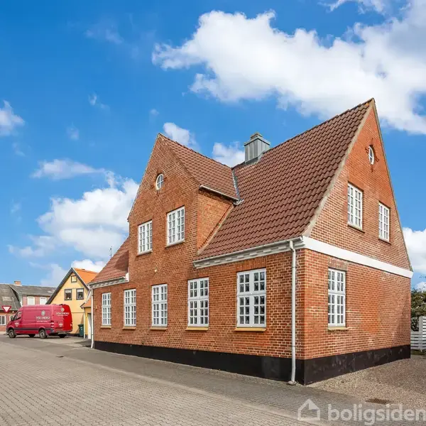 Et rødt murstenshus med hvide vinduer står på en brolagt gade under en blå, skyet himmel. I baggrunden ses flere bygninger og en rød varevogn.