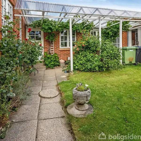 En have med en stenbelagt sti fører til en murstensbygning. En pergola dækket af grønne planter skaber en skyggefuld gang. Græsplæne med en dekorativ krukke til højre.